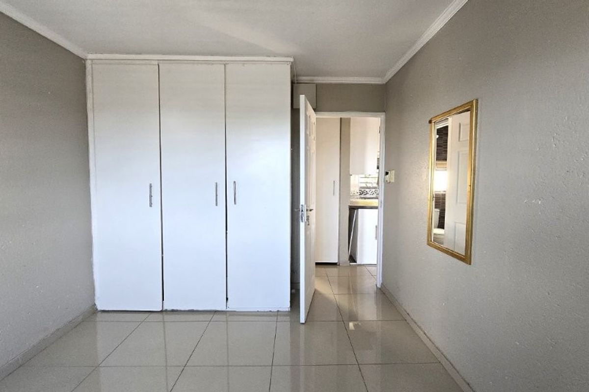 A vacant bedroom featuring neutral tiled flooring, built-in white cupboards, and coving, with a view through to an adjacent room.