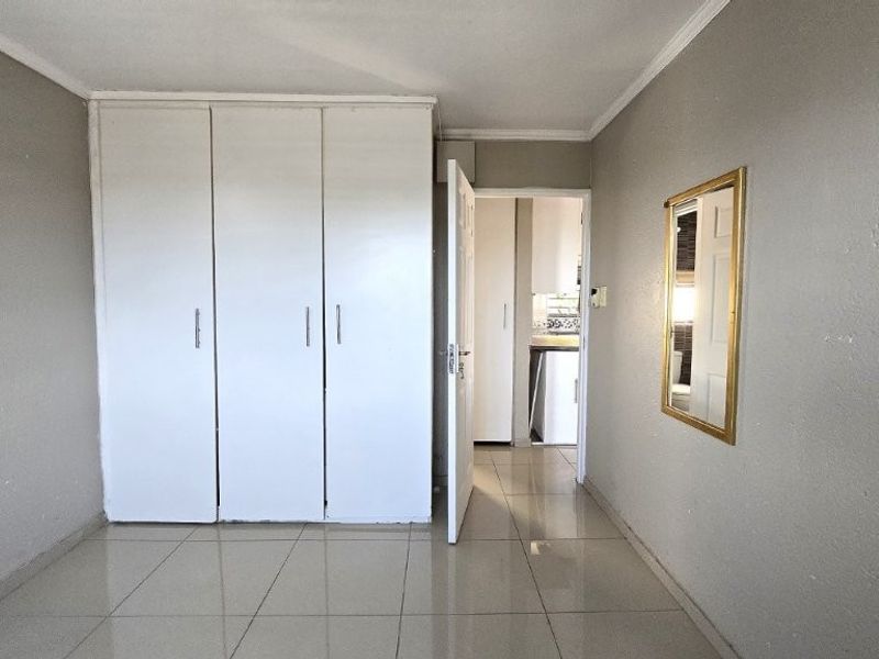 A vacant bedroom featuring neutral tiled flooring, built-in white cupboards, and coving, with a view through to an adjacent room.