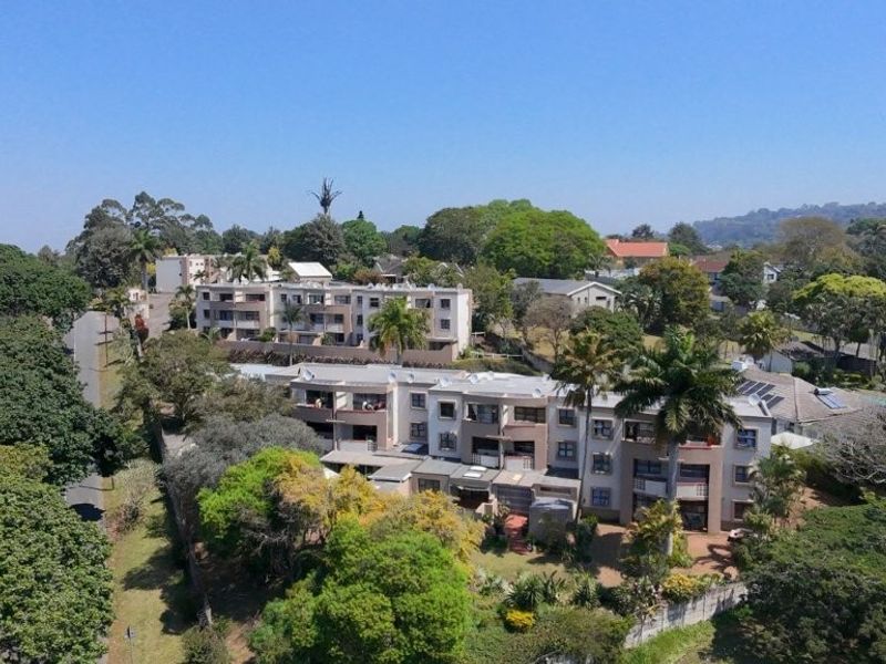 An aerial view of a multi-story townhouse complex set within a lush, green residential neighborhood with established trees and paved access.
