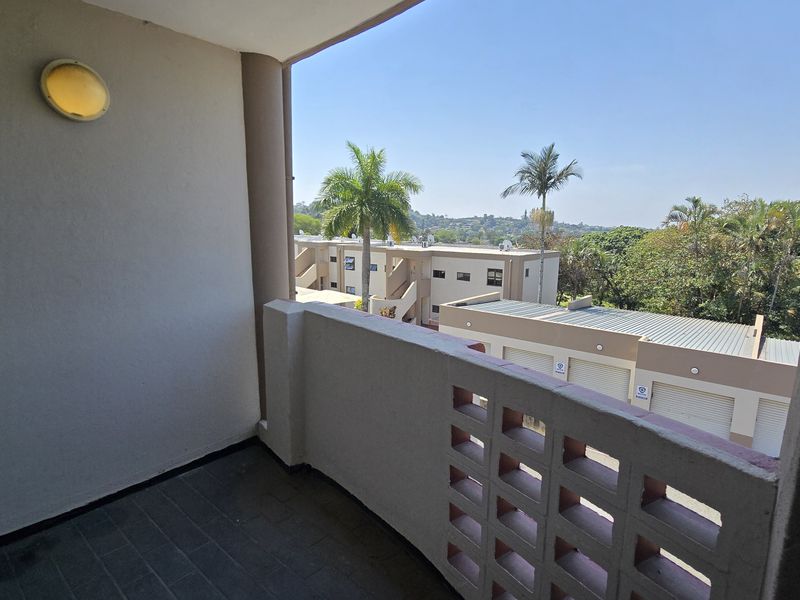 A covered balcony featuring tiled flooring, a wall-mounted light, and a decorative breeze block balustrade, offering an elevated view of the surrounding complex.
