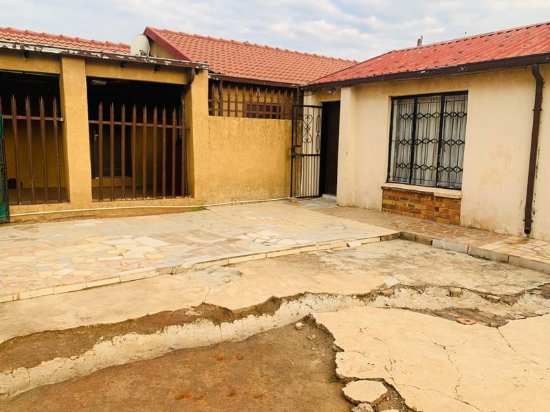 Rear exterior view of the property featuring a paved patio area, security gates, and a tiled roof, showing significant wear to the concrete surfaces.