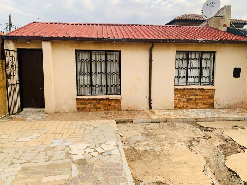 Rear exterior view of a single-story house featuring a corrugated metal roof, security-fitted windows, and a paved patio area requiring maintenance.