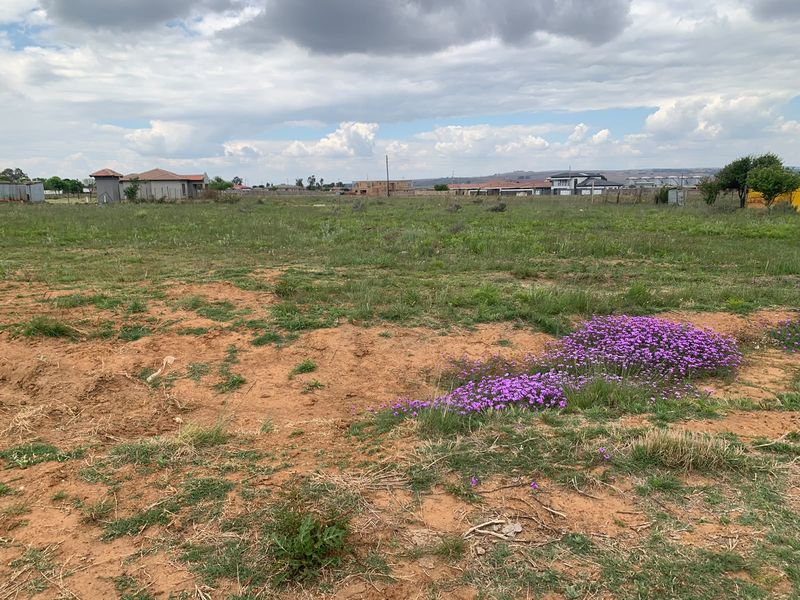 A wide-angle view of a vacant residential plot featuring natural ground cover and surrounding neighborhood development.