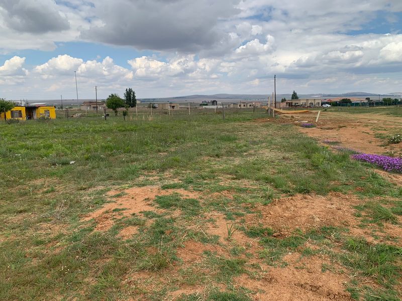 A wide-angle view of a vacant residential plot featuring natural grass, soil, wire fencing, and surrounding neighborhood infrastructure.