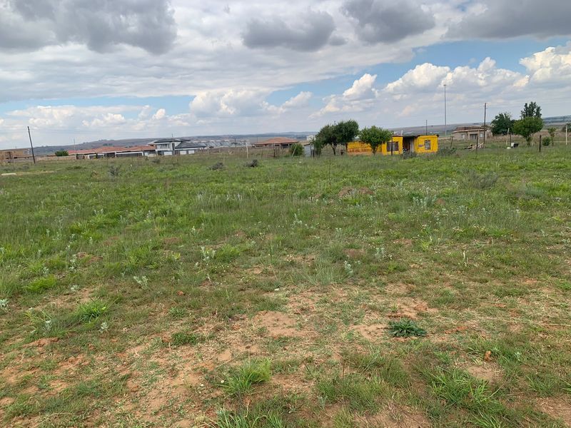 A wide-angle view of a vacant residential plot featuring natural grassland and perimeter fencing, situated in a developing suburban area.