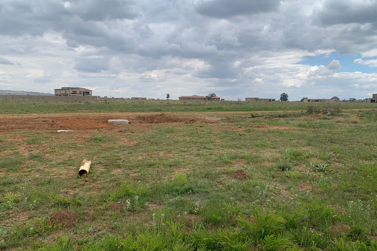 A vacant residential plot featuring natural grass coverage, an exposed drainage pipe, and a manhole cover, set within a developing residential estate.
