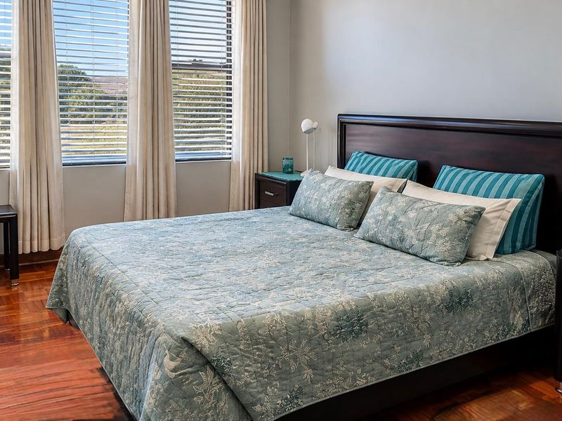 A well-maintained bedroom featuring warm parquet flooring and large windows with venetian blinds, offering a bright and comfortable living space.