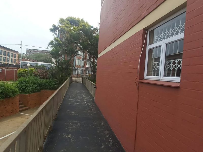 Exterior view of the flat showing a paved walkway alongside the brick building facade with security bars on the window.