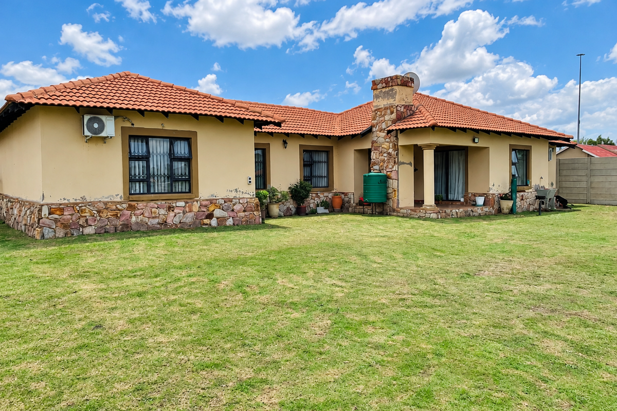 Rear view of a single-story house featuring a terracotta tile roof, stone cladding accents, and a spacious lawn area with a water tank.