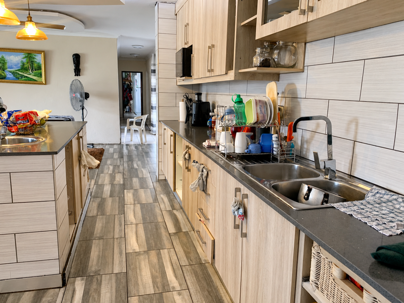 A modern kitchen featuring light wood-tone cabinetry, stone countertops, a double stainless steel sink, and contemporary tiled backsplash and flooring.