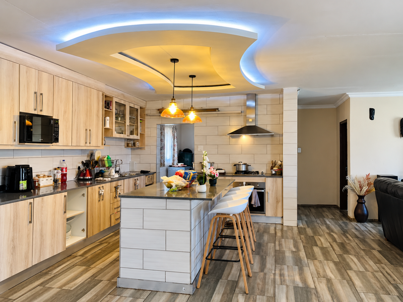 Modern open-plan kitchen featuring light wood cabinetry, a central island with seating, integrated appliances, and a distinctive illuminated recessed ceiling design.