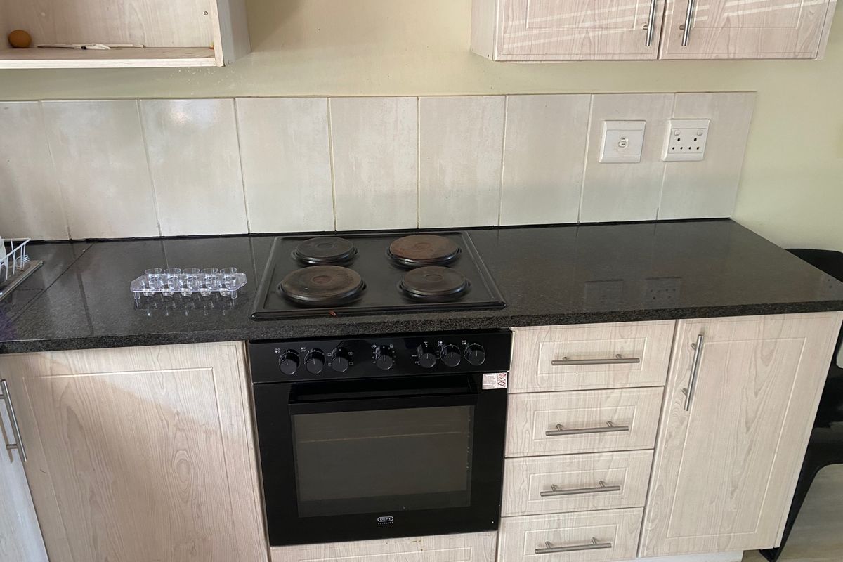 A functional kitchen featuring light wood-grain cabinetry, dark laminate countertops, a built-in electric oven, and a four-plate hob with a tiled backsplash.