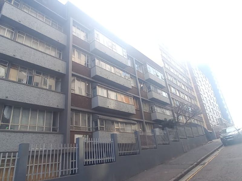Street-level view of a multi-story apartment building featuring a brick and concrete facade with integrated balconies and perimeter security fencing.