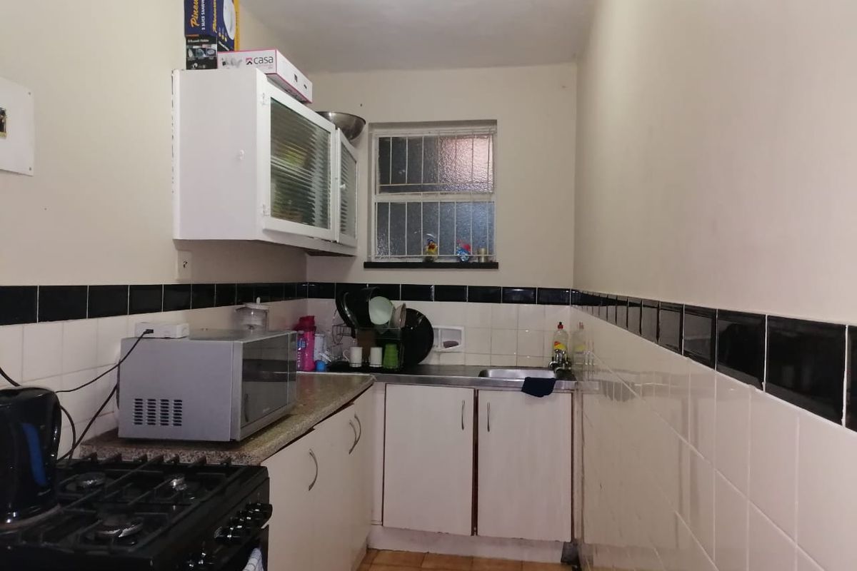 A compact kitchen featuring built-in cabinetry, a gas stove, and a stainless steel sink with tiled splashbacks and security bars on the window.