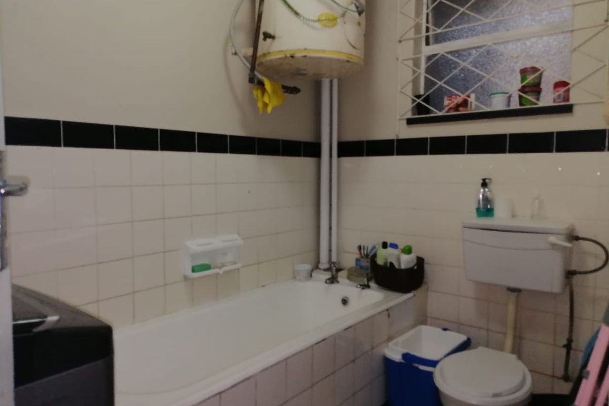 A functional bathroom featuring a standard bathtub, toilet, and wall-mounted geyser, with classic tiled wall finishes and security bars on the window.