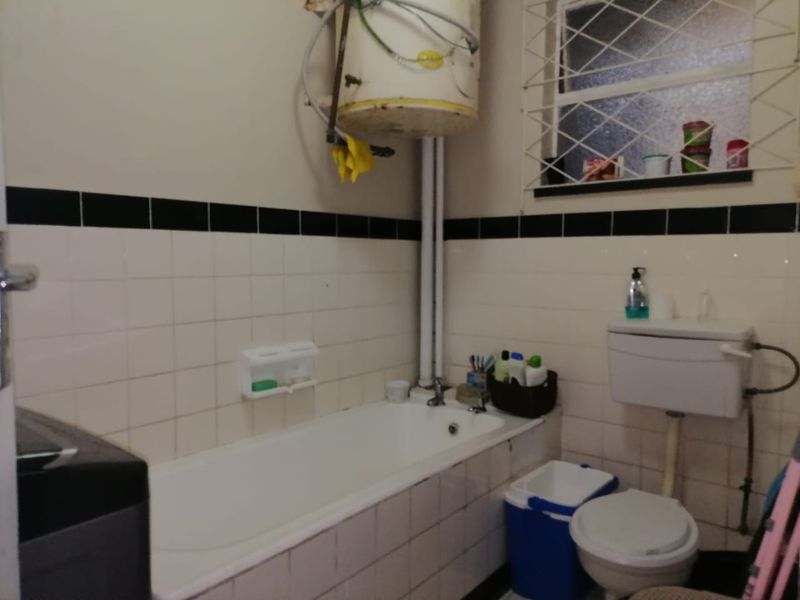 A functional bathroom featuring a standard bathtub, toilet, and wall-mounted geyser, with classic tiled wall finishes and security bars on the window.