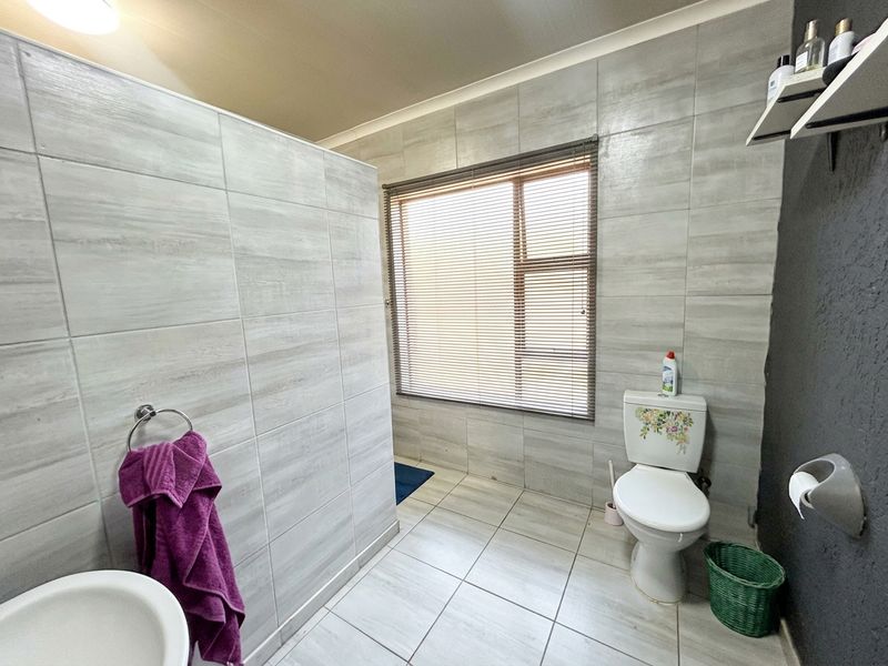 A functional bathroom featuring light grey wall and floor tiles, a window with fitted blinds, and standard sanitary fixtures.