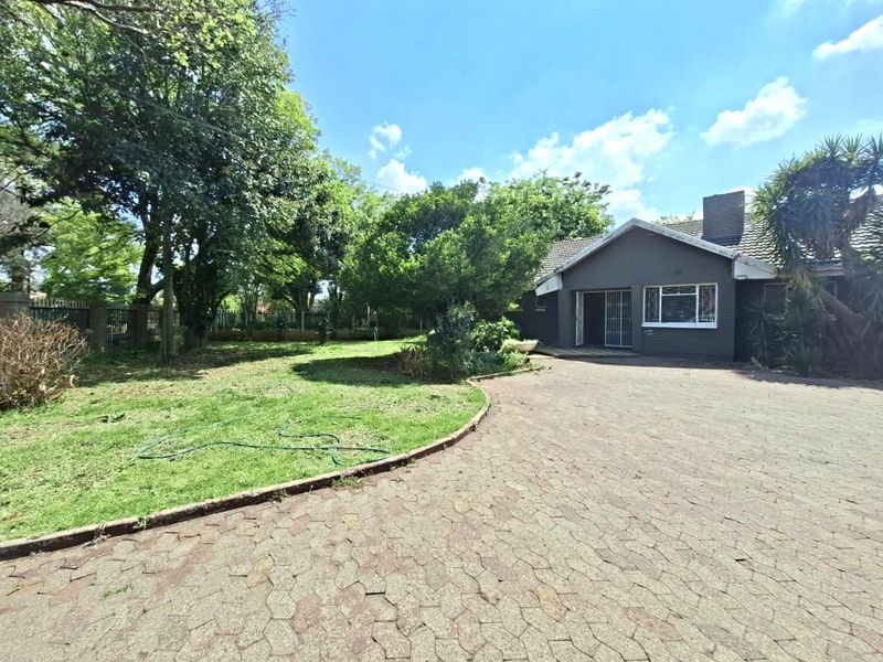 Front view of a single-story house featuring a paved driveway, brick exterior, and established garden with mature trees.