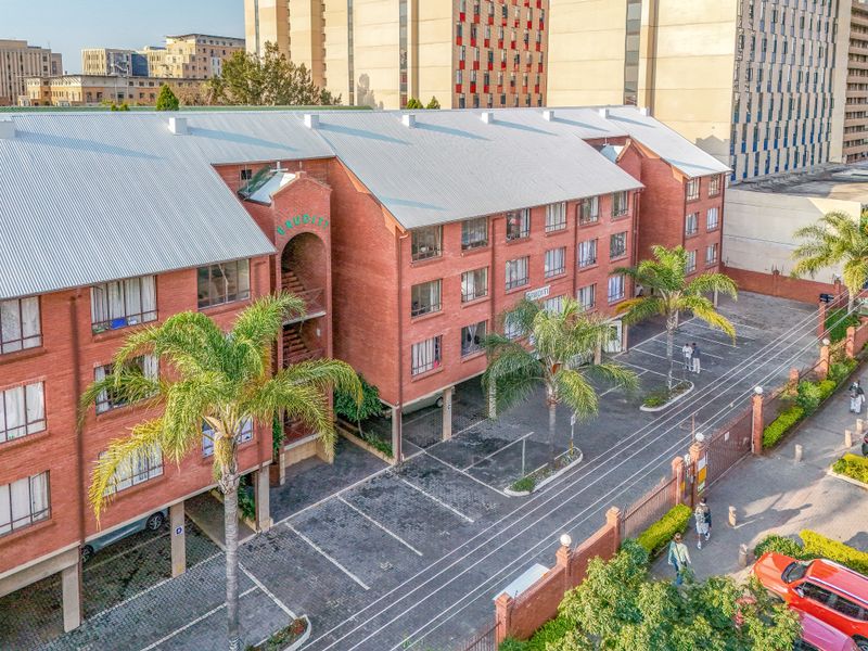 Aerial view of a multi-story brick apartment complex featuring a paved parking area, metal roofing, and secure perimeter fencing.