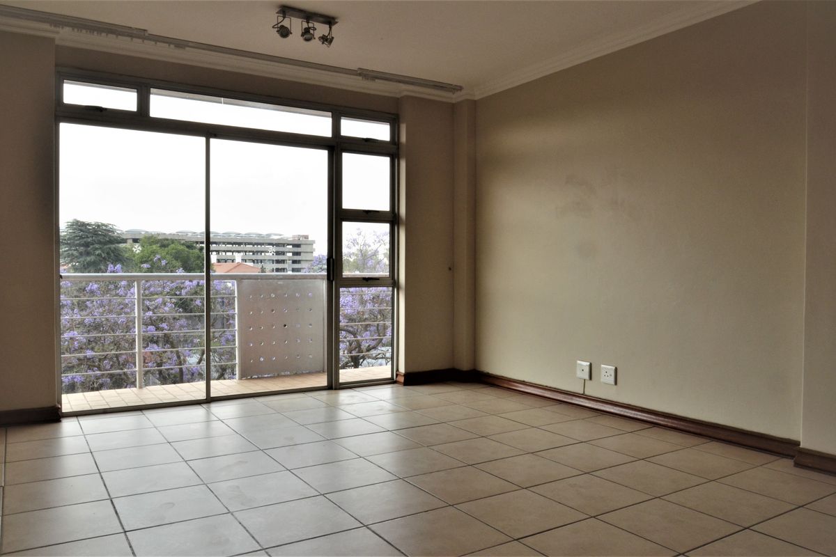 An empty, sunlit living area featuring neutral tiled flooring and a large sliding glass door that opens onto a private balcony.