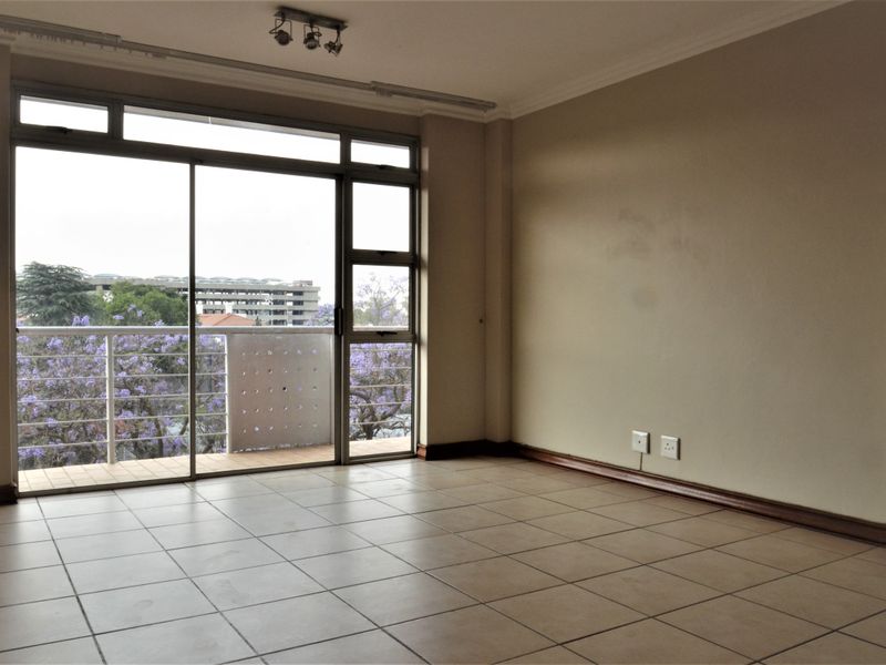 An empty, sunlit living area featuring neutral tiled flooring and a large sliding glass door that opens onto a private balcony.