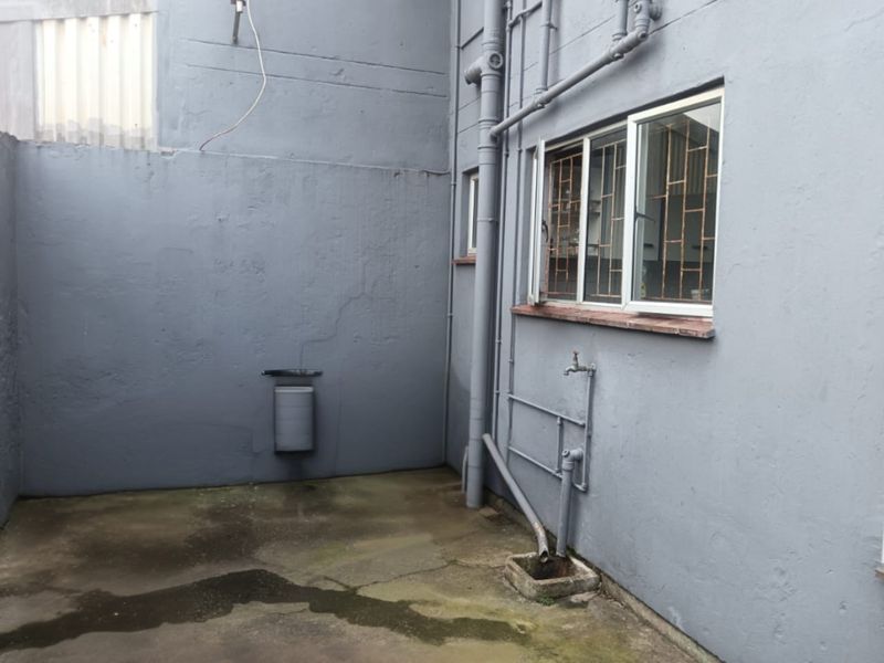 A rear exterior utility area featuring concrete paving, exposed drainage pipes, and a window with security bars.
