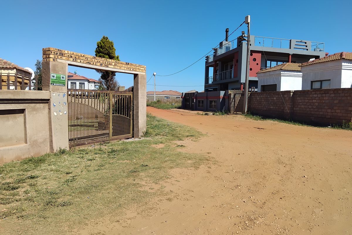 Street view showing the property entrance with a brick boundary wall, metal gate, and access to the multi-story residential dwelling via an unpaved road.