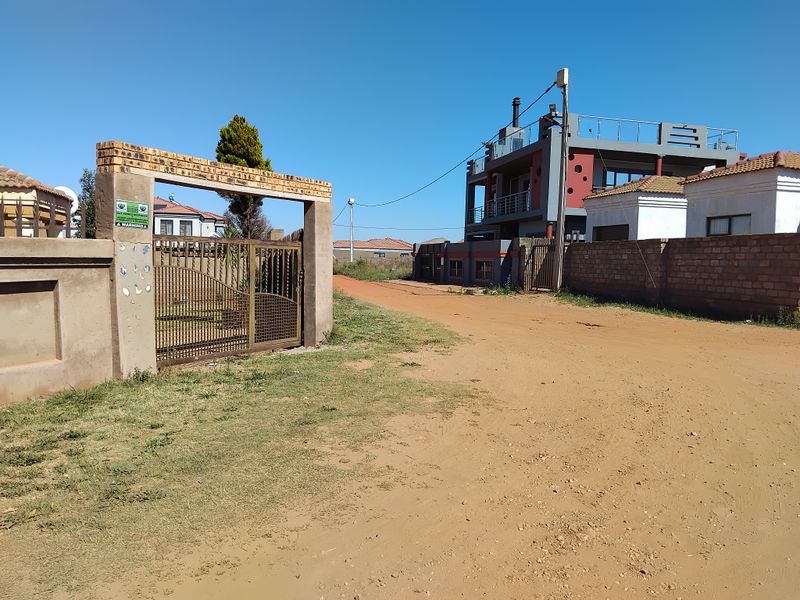 Street view showing the property entrance with a brick boundary wall, metal gate, and access to the multi-story residential dwelling via an unpaved road.