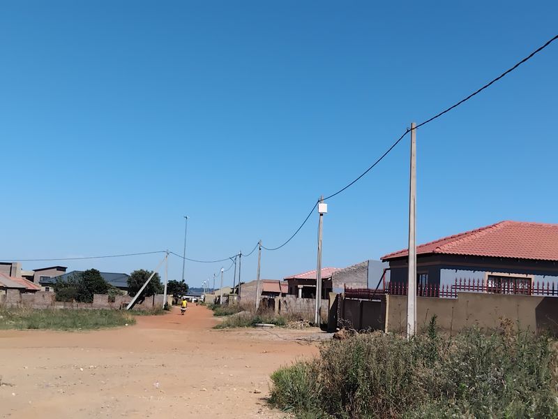A street view showing the neighbourhood approach with unpaved roads, overhead utility infrastructure, and residential properties with boundary walls.