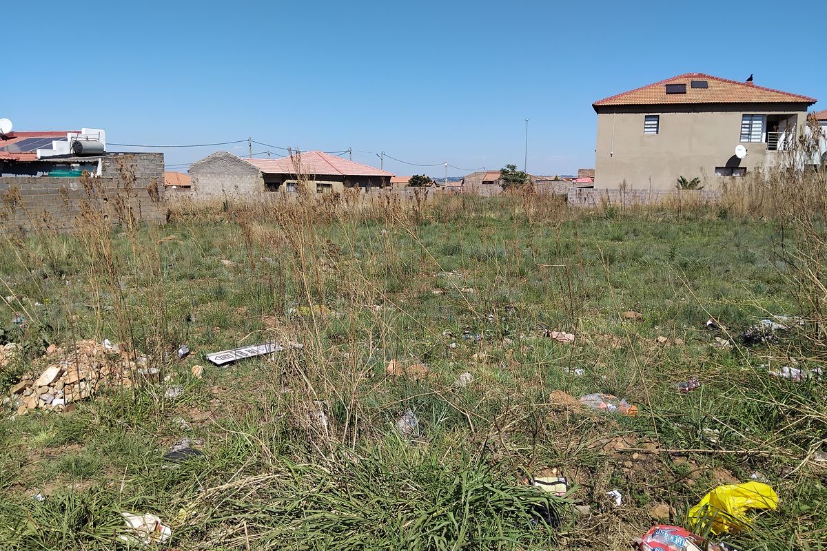 An undeveloped residential plot featuring overgrown grass and boundary walls, situated in a suburban neighborhood setting.