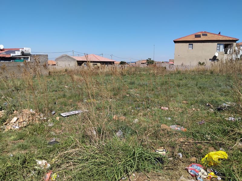 An undeveloped residential plot featuring overgrown grass and boundary walls, situated in a suburban neighborhood setting.