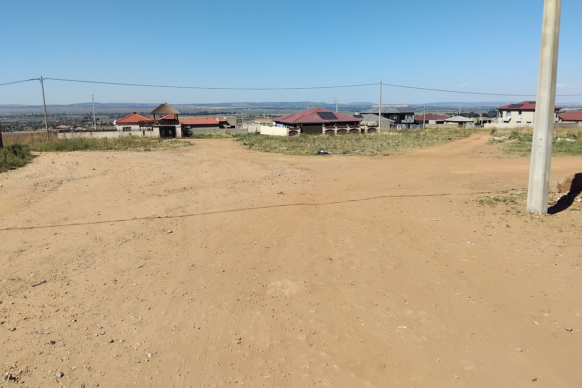 A view of an unpaved road and utility infrastructure in a residential area with surrounding houses in the distance.