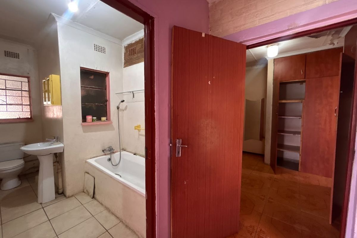 A dated bathroom featuring a pedestal basin and built-in bathtub, adjacent to a room with built-in wooden cupboards and tiled flooring.