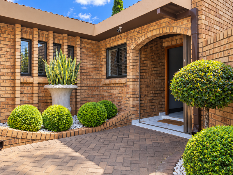 A well-maintained townhouse entrance featuring a face brick facade, herringbone paved driveway, and a secure, tiled entryway with an arched architectural detail.
