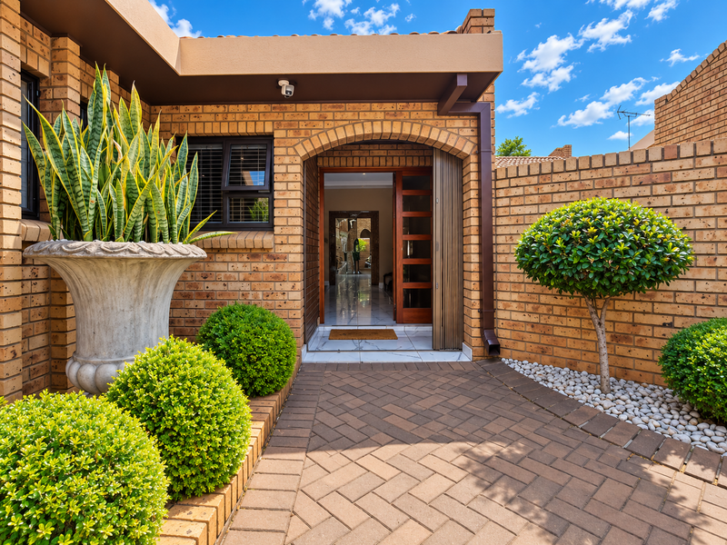 A well-maintained townhouse entrance featuring a face brick facade, arched doorway with timber-framed glass door, and a neat herringbone paved walkway.