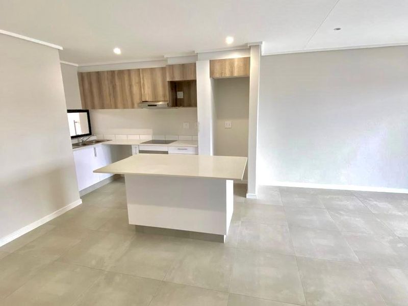 Modern open-plan kitchen featuring light wood-tone cabinetry, a central island with stone countertop, built-in hob, and neutral tiled flooring throughout the living space.