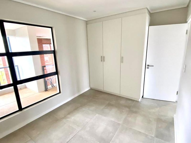 A bright, empty bedroom featuring modern large-format floor tiles, built-in cupboards, and a large window providing natural light.