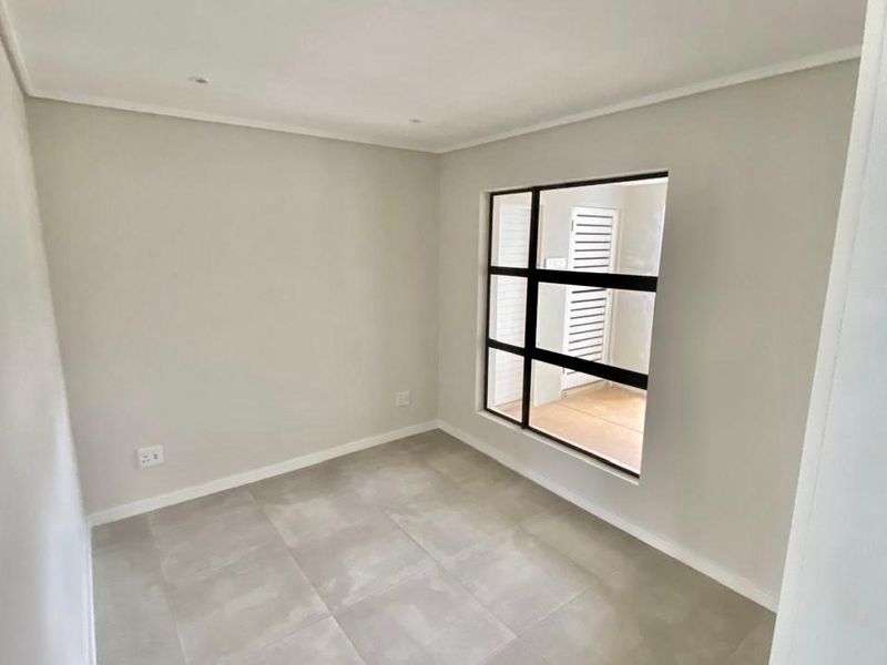 A bright, empty bedroom featuring modern large-format floor tiles, recessed downlighting, and a large black-framed window looking out onto a balcony.