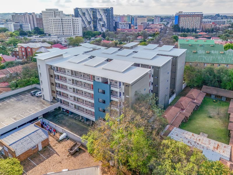 Aerial view of a multi-story apartment complex featuring a metal roof, exterior balconies, and an adjacent asphalt parking area within an urban setting.