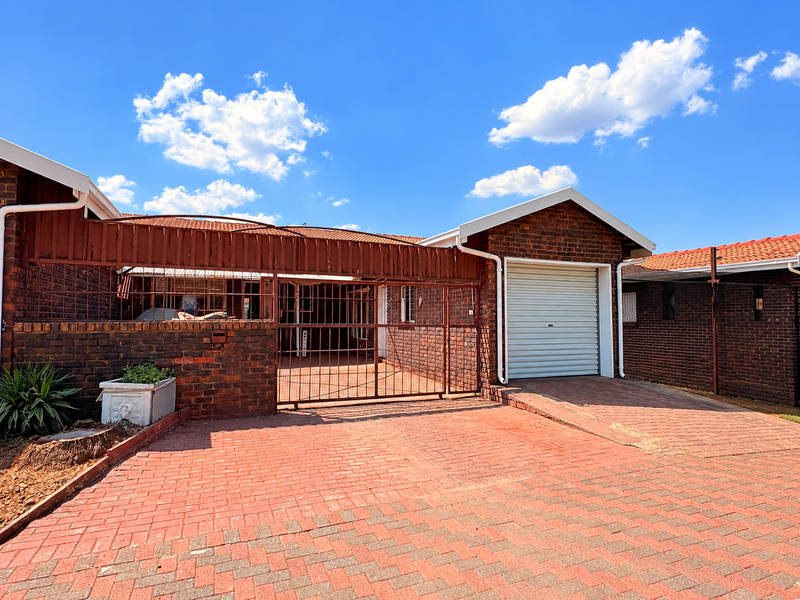 Front exterior view of a brick townhouse featuring a paved driveway, a single garage, and secure metal gate access.
