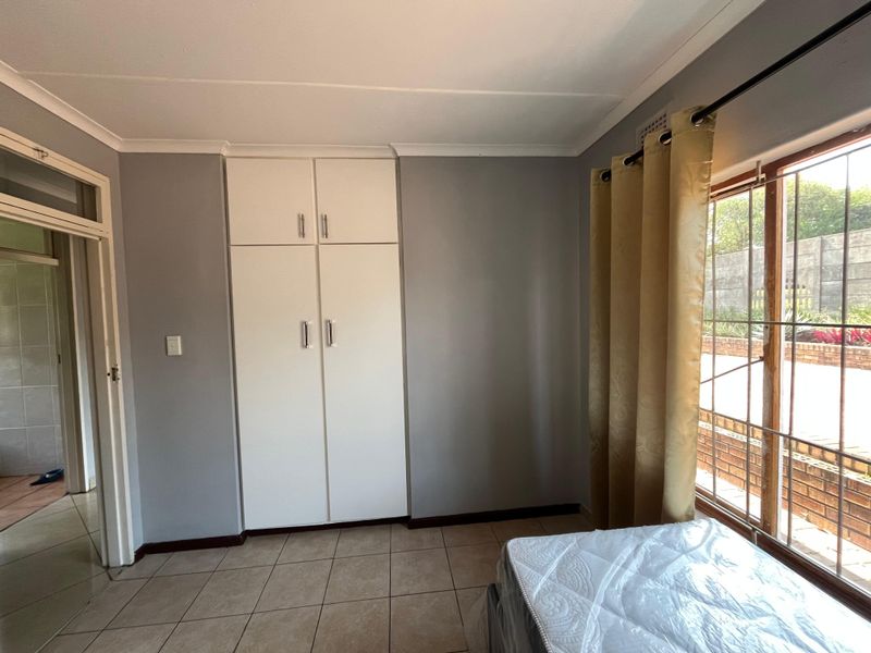 A bedroom featuring built-in cupboards, tiled flooring, and a window with security bars, offering a functional and neutral living space.