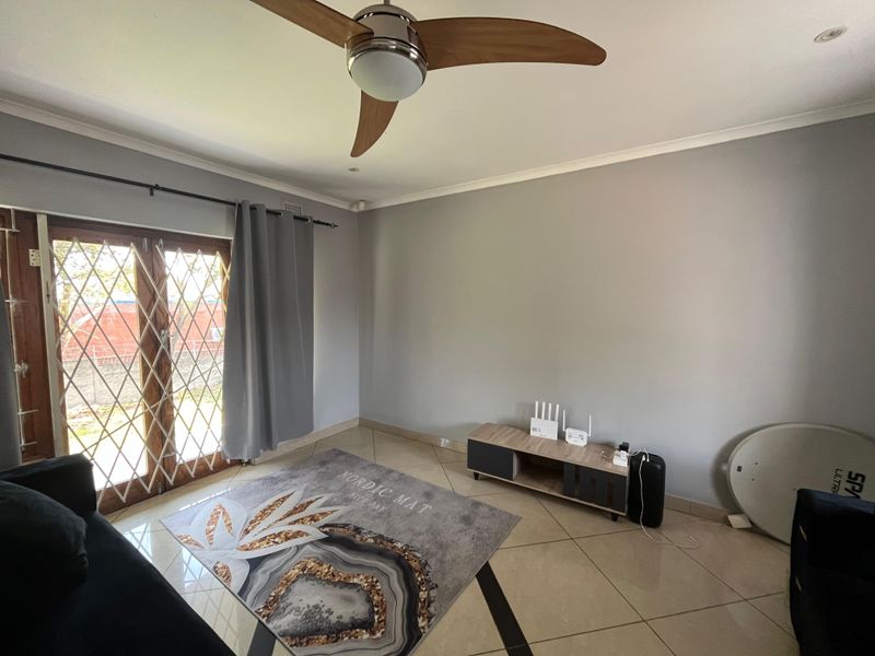 A bright living room featuring tiled flooring, a modern ceiling fan, and double doors with security gates leading to the exterior.