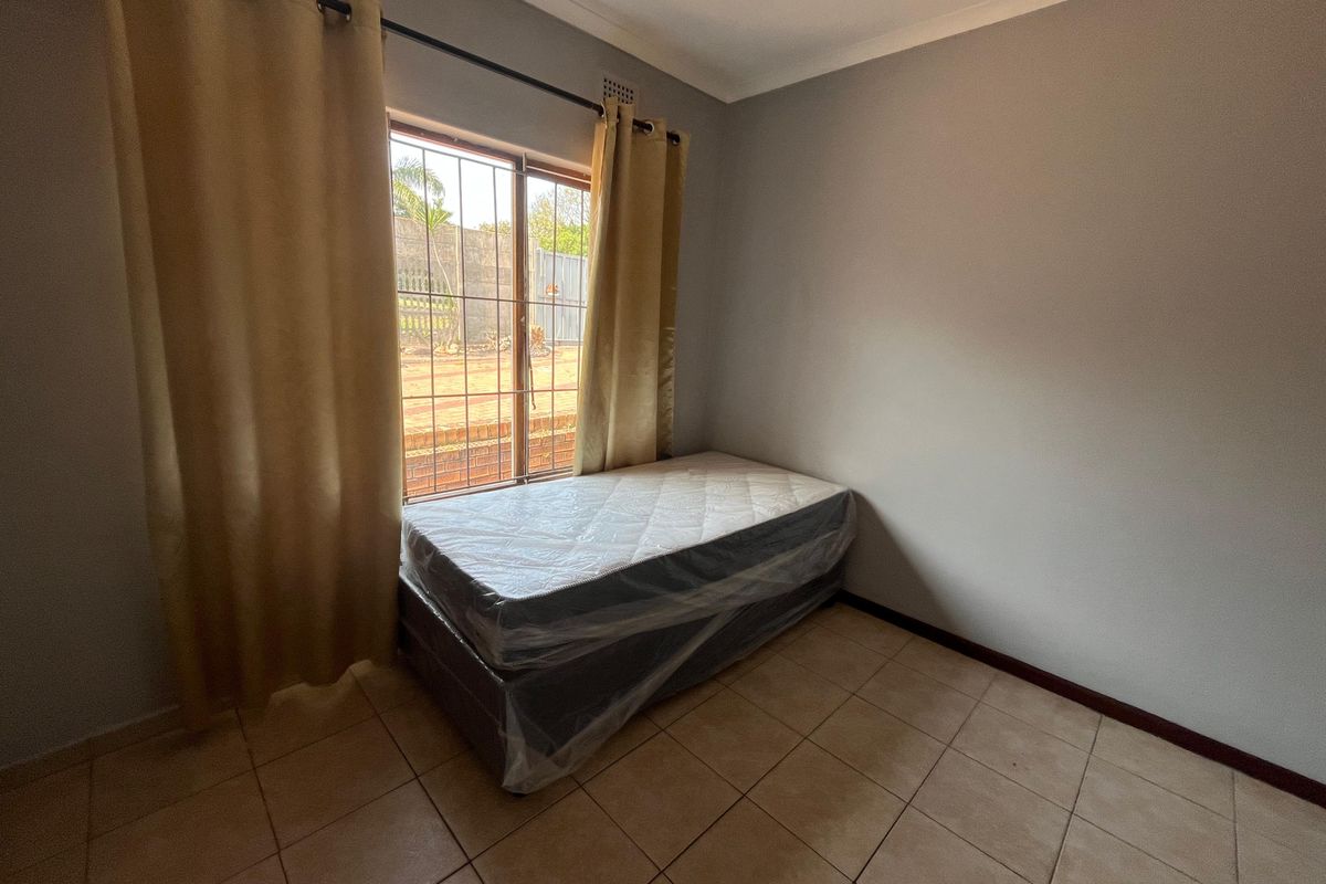 A simple bedroom featuring tiled flooring, neutral walls, and a window fitted with security bars.