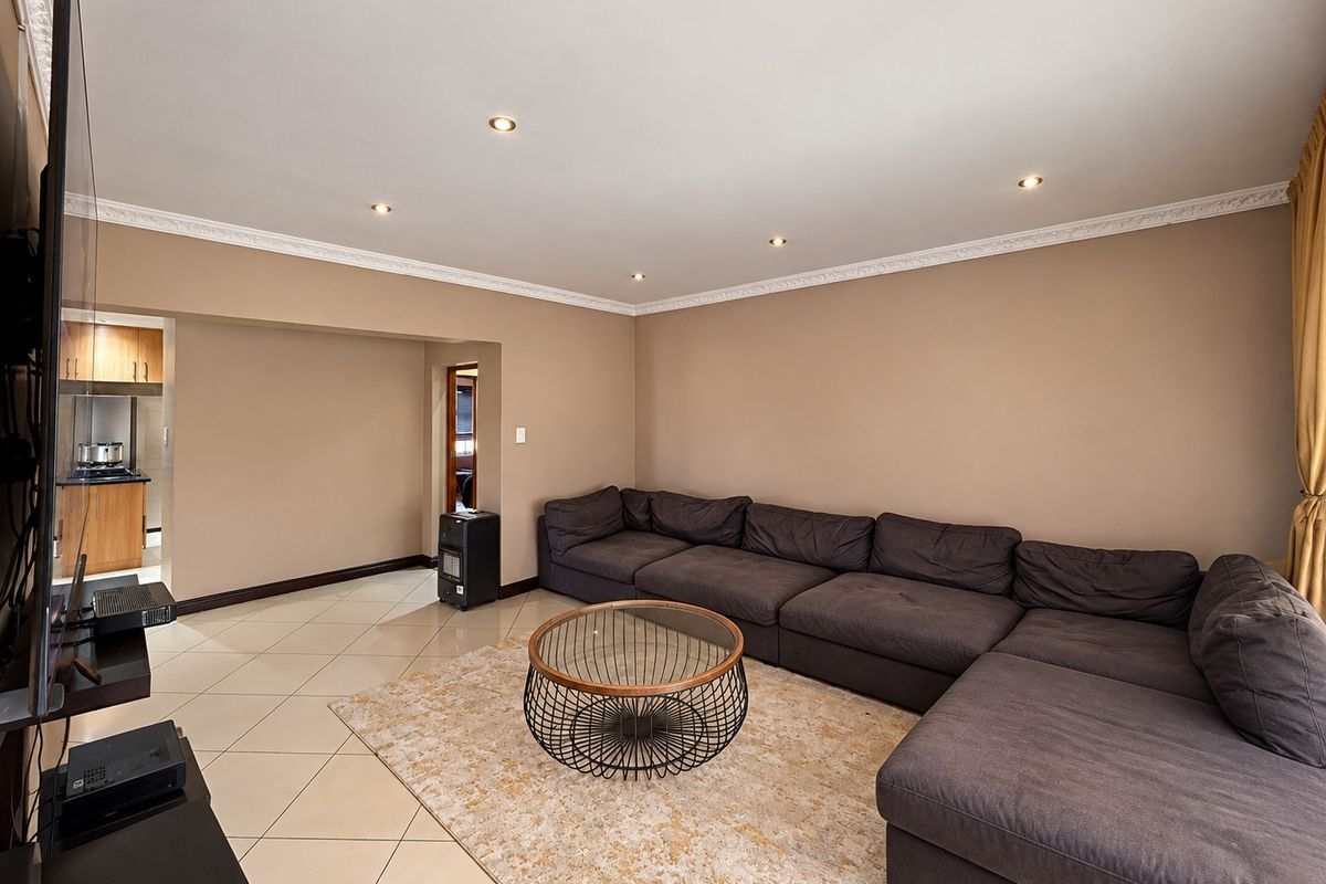 Spacious living room featuring neutral wall tones, tiled flooring, recessed downlighting, and decorative ceiling cornicing, with an open-plan flow to the adjacent kitchen area.