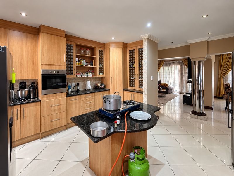 Open plan kitchen featuring wooden cabinetry, granite countertops, and built-in oven, flowing into a spacious living area with tiled flooring and decorative pillar.