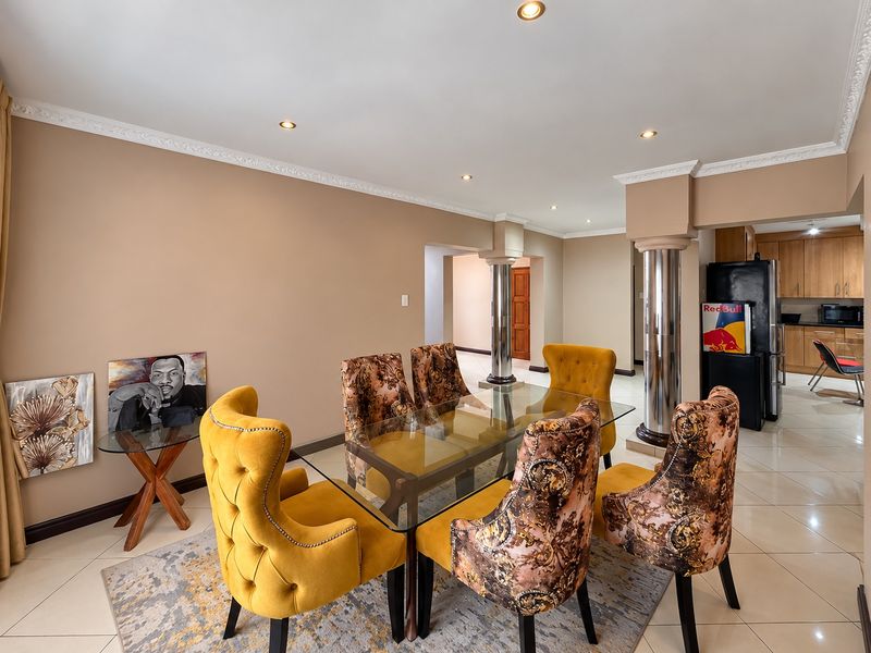 Open-plan dining area featuring decorative ceiling cornices, recessed lighting, and chrome columns, seamlessly flowing into a functional kitchen space with tiled flooring.