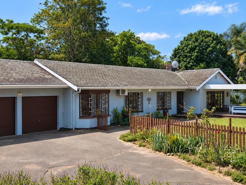 A well-maintained single-story house featuring a double garage, paved driveway, wooden picket fencing, and a covered patio area set within a landscaped garden.