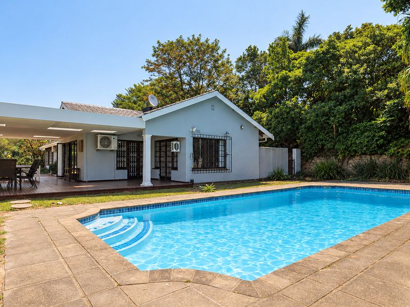 A well-maintained outdoor entertainment area featuring a swimming pool, a spacious covered patio with decking, and a built-in stone fireplace in the garden.