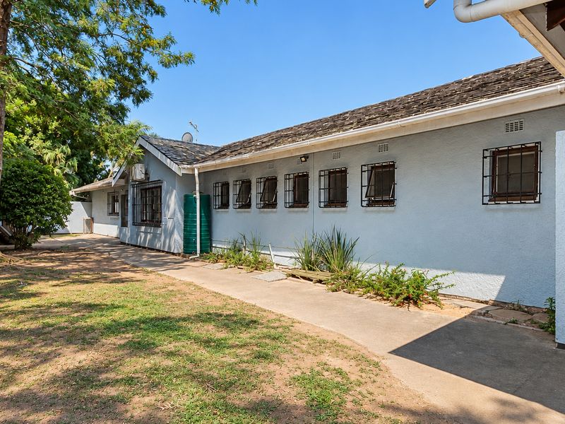 Rear exterior view of the house featuring a shingle roof, security-barred windows, a water tank, and a concrete pathway alongside a stone retaining wall.