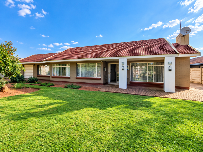 A well-maintained single-story house featuring a tiled roof, brick facade, and a spacious front lawn with a paved patio area.